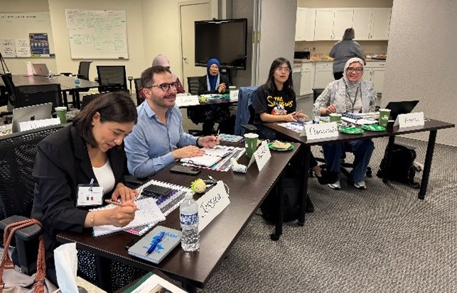 A classroom setting with several participants seated at tables during a training session. They are taking notes, looking at materials and laptops, and have name cards in front of them. The room includes whiteboards, a large monitor and shelves in the background.